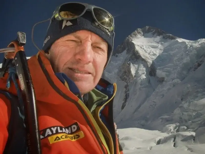 A middle-aged mountaineer takes a selfie in high-altitude conditions, squinting and smiling slightly against bright sunlight, wearing a dark beanie, glacier sunglasses pushed up on his forehead, and a bright orange down jacket with "KAYLAND" and "ACERBIS" sponsor logos on the chest. An ice axe is strapped to his backpack over his left shoulder, and the iconic snow-covered peak of Mount Everest looms prominently in the background under a clear blue sky.