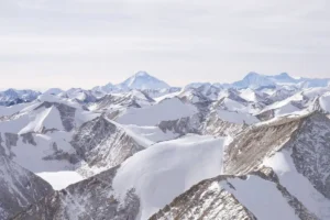A breathtaking aerial or distant panoramic view of the snow-covered Himalayan mountain range in Nepal, with rugged rocky ridges and peaks partially blanketed in snow in the foreground, leading to the prominent pyramid-shaped summit of Mount Everest rising prominently in the background under a pale, overcast sky.