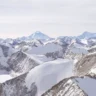 A breathtaking aerial or distant panoramic view of the snow-covered Himalayan mountain range in Nepal, with rugged rocky ridges and peaks partially blanketed in snow in the foreground, leading to the prominent pyramid-shaped summit of Mount Everest rising prominently in the background under a pale, overcast sky.