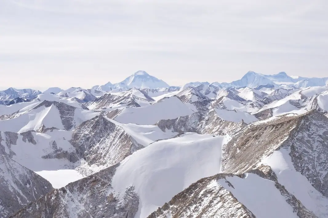 A breathtaking aerial or distant panoramic view of the snow-covered Himalayan mountain range in Nepal, with rugged rocky ridges and peaks partially blanketed in snow in the foreground, leading to the prominent pyramid-shaped summit of Mount Everest rising prominently in the background under a pale, overcast sky.