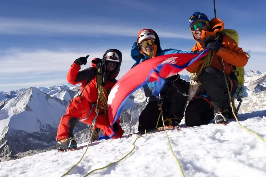 Three mountaineers in full high-altitude climbing gear, including helmets, goggles, oxygen masks, and insulated suits, celebrate on a snowy mountain summit under a clear blue sky; they are roped together for safety, posing joyfully while holding and displaying a large Nepali national flag with its distinctive sun and moon symbols