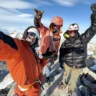 Three mountaineers celebrate on a snowy alpine summit, taking a selfie against a stunning backdrop of jagged peaks and blue sky. The person on the left, in an orange jacket, holds the camera while making a shaka hand gesture. The middle climber, also in orange, flashes a big smile, and the climber on the right, wearing a black puffy jacket and white helmet, raises a triumphant fist. All are equipped with climbing gear, helmets, and sunglasses, standing on rocky, snow-dusted ground high above the clouds.