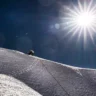 A lone mountaineer in full high-altitude gear climbs a steep, snow-covered ridge under a deep blue sky. The bright sun dominates the upper right, creating intense lens flares and starburst effects, while a fixed rope trails down the slope behind the climber, emphasizing the extreme exposure and altitude.