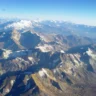 Aerial view of a vast, rugged mountain range under a clear blue sky. Sharp, jagged peaks with patches of snow dominate the foreground and midground, while distant snow-capped summits fade into the horizon against a bright, cloudless sky.