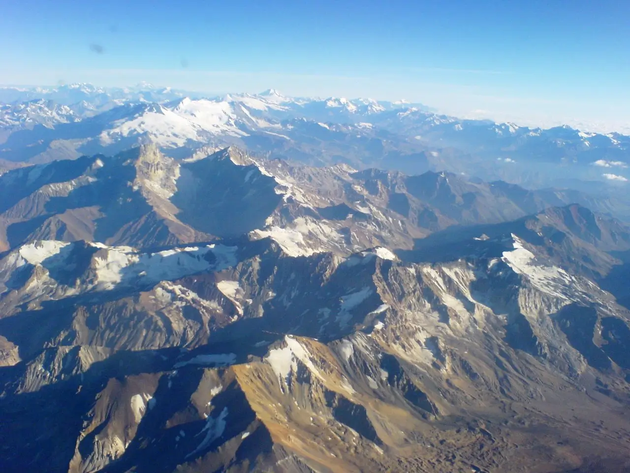 Aerial view of a vast, rugged mountain range under a clear blue sky. Sharp, jagged peaks with patches of snow dominate the foreground and midground, while distant snow-capped summits fade into the horizon against a bright, cloudless sky.