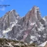 A dramatic view of a jagged, snow-patched alpine peak in the Caucasus Mountains under a clear blue sky. Colored lines (yellow for 2025, red for 2024, blue for 2016) trace different climbing routes up the steep rock face. Georgian text in the top left mentions the peak "Ushba," its elevation of 4690 m (north summit) and 4710 m (south summit), and route grades M5+, 5B (Caucasian), with a difficulty of 680 m.