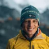 A smiling mountaineer poses for a close-up portrait in a snowy alpine environment, looking directly at the camera with a relaxed expression. He wears a teal La Sportiva beanie with patches including "VP Bank" and "La Sportiva," an orange neck gaiter, and a bright yellow La Sportiva jacket. The blurred background shows a misty, snow-covered mountainous landscape.