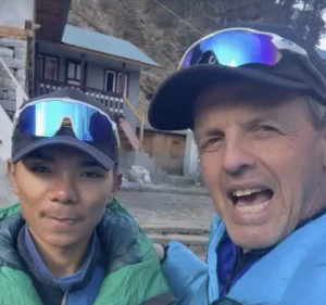 Two people pose for a selfie in a mountainous village setting. The person on the left, wearing a green puffy jacket and reflective blue ski goggles on their cap, looks calmly at the camera. The person on the right, in a bright blue jacket with reflective goggles pushed up on their cap, has their arm around the other and is grinning widely. Behind them is a stone and timber guesthouse with a green roof, concrete steps, and a rocky hillside with sparse trees.
