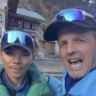 Two people pose for a selfie in a mountainous village setting. The person on the left, wearing a green puffy jacket and reflective blue ski goggles on their cap, looks calmly at the camera. The person on the right, in a bright blue jacket with reflective goggles pushed up on their cap, has their arm around the other and is grinning widely. Behind them is a stone and timber guesthouse with a green roof, concrete steps, and a rocky hillside with sparse trees.