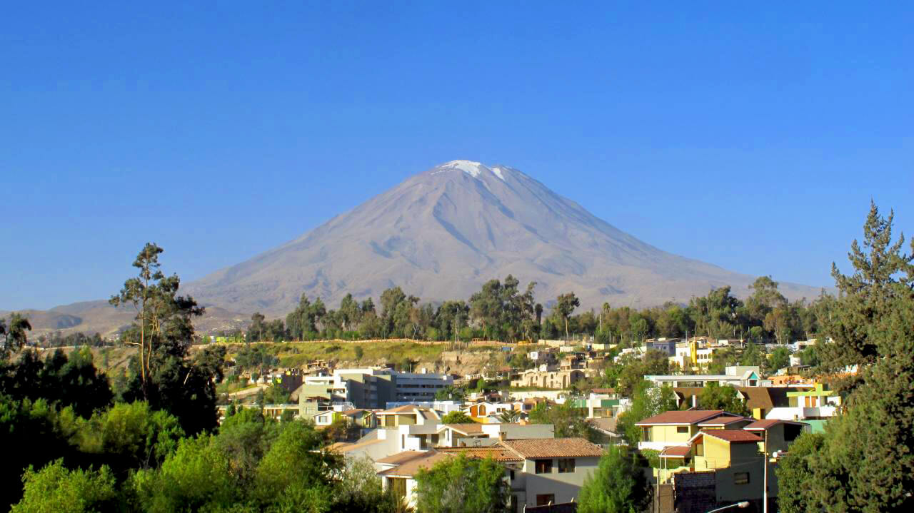 A perfectly symmetrical, snow-capped stratovolcano towers majestically against a clear, deep blue sky. In the foreground, a residential neighborhood with colorful houses, green trees, and lush vegetation spreads across the hillside, creating a striking contrast between the urban settlement and the massive, serene volcanic peak that dominates the background.