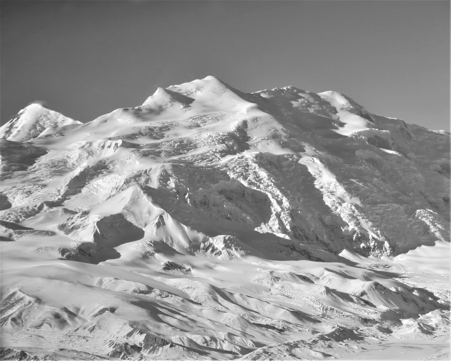 A striking black-and-white aerial photograph of a dramatic, snow-covered alpine mountain range. Jagged peaks rise sharply, with deep shadowed valleys and extensive glaciers flowing between them, their crevassed surfaces catching the light. The foreground features a vast expanse of undulating snow and ice, creating a timeless, high-contrast glacial landscape under a clear sky.