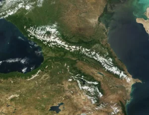 Satellite image of the Caucasus Mountains region, showing the prominent snow-capped Greater Caucasus range running horizontally across the center. The Black Sea is visible on the left, the Caspian Sea on the right, with greener terrain to the north and more arid land to the south.