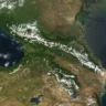 Satellite image of the Caucasus Mountains region, showing the prominent snow-capped Greater Caucasus range running horizontally across the center. The Black Sea is visible on the left, the Caspian Sea on the right, with greener terrain to the north and more arid land to the south.