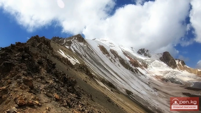 Rugged rocky slope with patches of snow ascending to towering snow-capped peaks in the high Pamir Mountains under a bright blue sky with scattered clouds