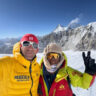 Two smiling mountaineers in bright expedition gear taking a selfie on a snowy glacier with massive ice-covered peaks in the background under a clear blue sky