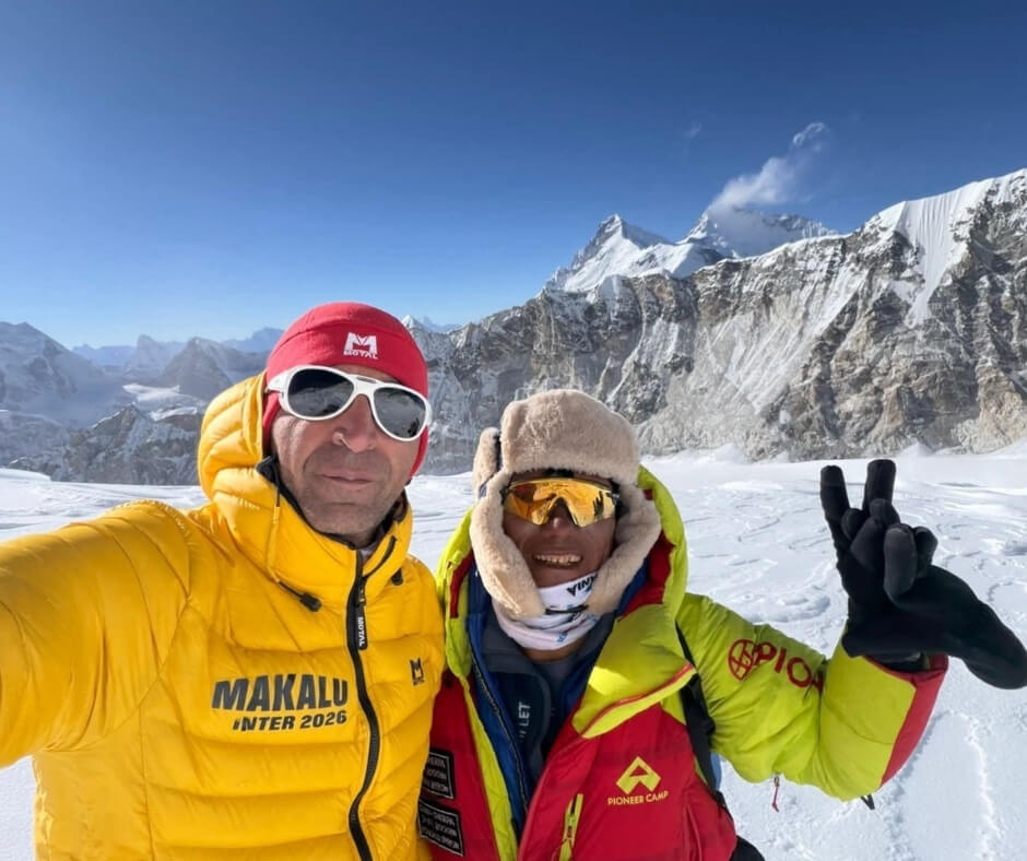 Two smiling mountaineers in bright expedition gear taking a selfie on a snowy glacier with massive ice-covered peaks in the background under a clear blue sky