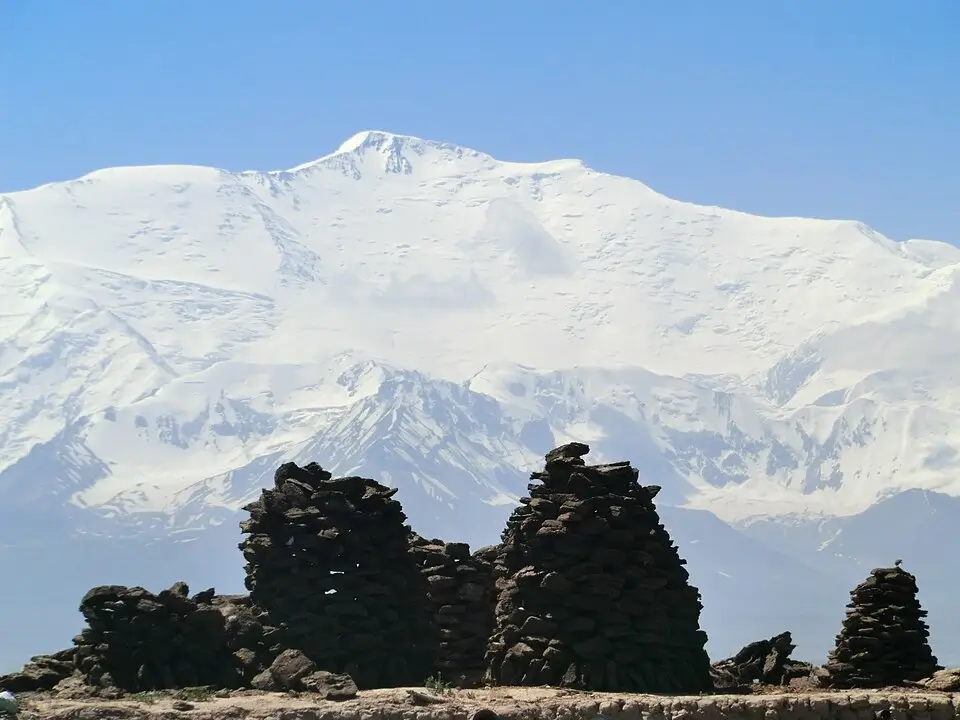 Traditional stone cairns or rock piles in the foreground with a massive snow-covered peak in the Pamir Mountains under a clear blue sky