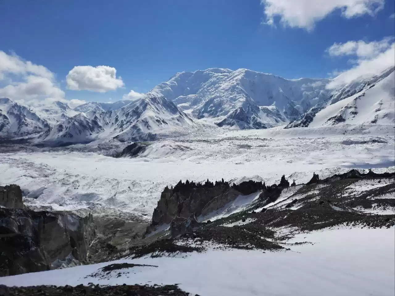 Expansive snow-covered glacier valley in the high Pamir Mountains, surrounded by rugged, snow-capped peaks under a bright blue sky with scattered clouds