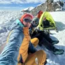 A group of three mountaineers taking a selfie on a snowy high-altitude glacier. Brightly colored tents are pitched behind them on the snow, with rugged peaks and blue sky in the background.