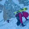 Two backcountry skiers ascending a steep, snowy alpine face in winter conditions. One skier in a red jacket climbs with ice axe and skis on their pack in the foreground, while another in blue follows higher up the icy slope, surrounded by dramatic rocky peaks and deep snow.