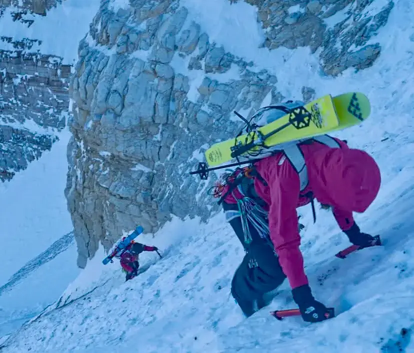 Two backcountry skiers ascending a steep, snowy alpine face in winter conditions. One skier in a red jacket climbs with ice axe and skis on their pack in the foreground, while another in blue follows higher up the icy slope, surrounded by dramatic rocky peaks and deep snow.