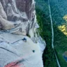Aerial view of a rock climber in blue on a vast smooth granite slab high on a dome, with red ropes leading down to another climber below; a narrow road winds through dense green forest far beneath, beside a small blue pond.