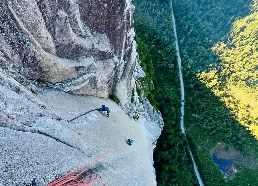 Aerial view of a rock climber in blue on a vast smooth granite slab high on a dome, with red ropes leading down to another climber below; a narrow road winds through dense green forest far beneath, beside a small blue pond.
