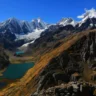 A breathtaking panoramic landscape of the Peruvian Andes, likely in the Cordillera Huayhuash or nearby range. Towering, jagged snow-capped peaks and glaciers dominate the horizon under a vibrant clear blue sky with a few wispy clouds. In the foreground and midground, rugged rocky terrain with brown and orange-hued slopes descends into a deep valley.