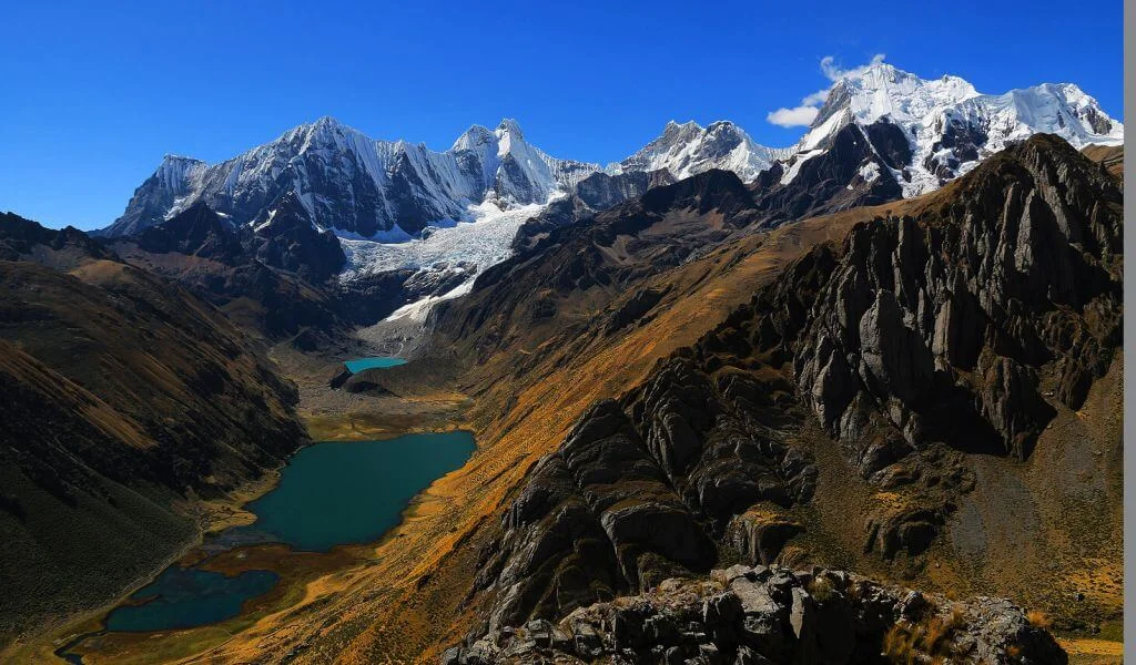 A breathtaking panoramic landscape of the Peruvian Andes, likely in the Cordillera Huayhuash or nearby range. Towering, jagged snow-capped peaks and glaciers dominate the horizon under a vibrant clear blue sky with a few wispy clouds. In the foreground and midground, rugged rocky terrain with brown and orange-hued slopes descends into a deep valley.