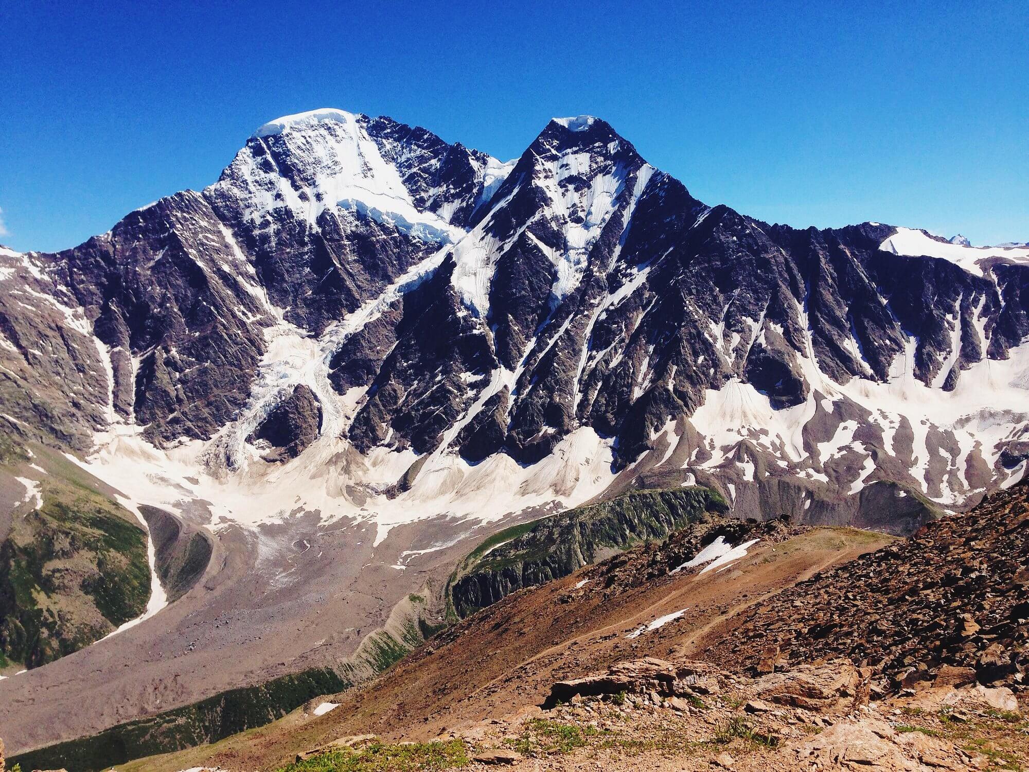 Two sharp, jagged twin mountain peaks heavily covered in snow and ice, with steep glaciers cascading down their faces. The foreground shows rocky alpine terrain with patches of green vegetation under a deep blue sky.
