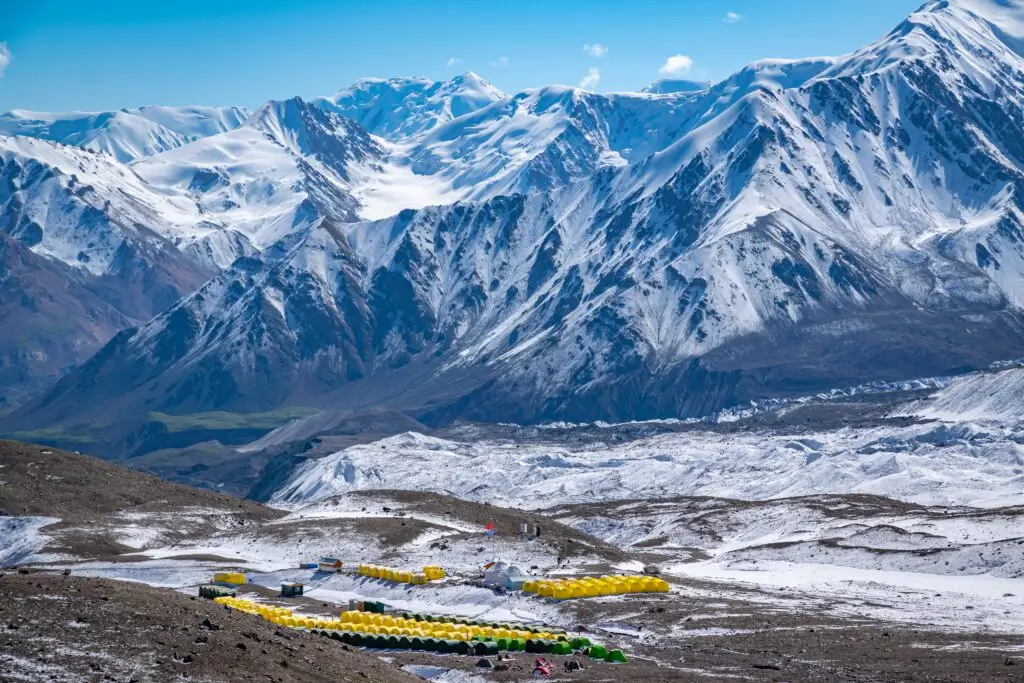 Colorful yellow and green tents of a mountaineering base camp set against towering snow-covered peaks in the Pamir Mountains under a clear blue sky
