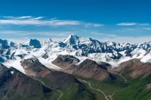 Aerial view of snow-capped peaks and massive glaciers in a rugged high mountain range under a clear blue sky, with rocky ridges, ice fields, and green valleys below.