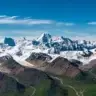Aerial view of snow-capped peaks and massive glaciers in a rugged high mountain range under a clear blue sky, with rocky ridges, ice fields, and green valleys below.