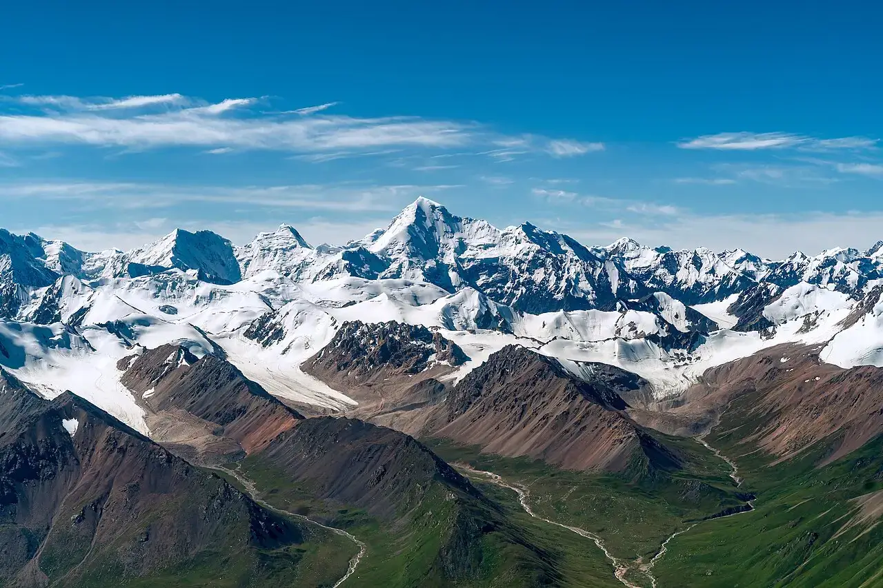Aerial view of snow-capped peaks and massive glaciers in a rugged high mountain range under a clear blue sky, with rocky ridges, ice fields, and green valleys below.