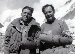 Historic black-and-white photo of Italian climbers Achille Compagnoni (left) and Lino Lacedelli (right) during the 1954 first ascent of K2. They stand arm-in-arm on a rocky, snowy slope with mountain peaks in the background; Compagnoni wears a quilted down jacket with a serious expression, while the bearded Lacedelli smiles slightly in a wool sweater with a squirrel patch and thick mittens.