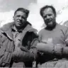 Historic black-and-white photo of Italian climbers Achille Compagnoni (left) and Lino Lacedelli (right) during the 1954 first ascent of K2. They stand arm-in-arm on a rocky, snowy slope with mountain peaks in the background; Compagnoni wears a quilted down jacket with a serious expression, while the bearded Lacedelli smiles slightly in a wool sweater with a squirrel patch and thick mittens.