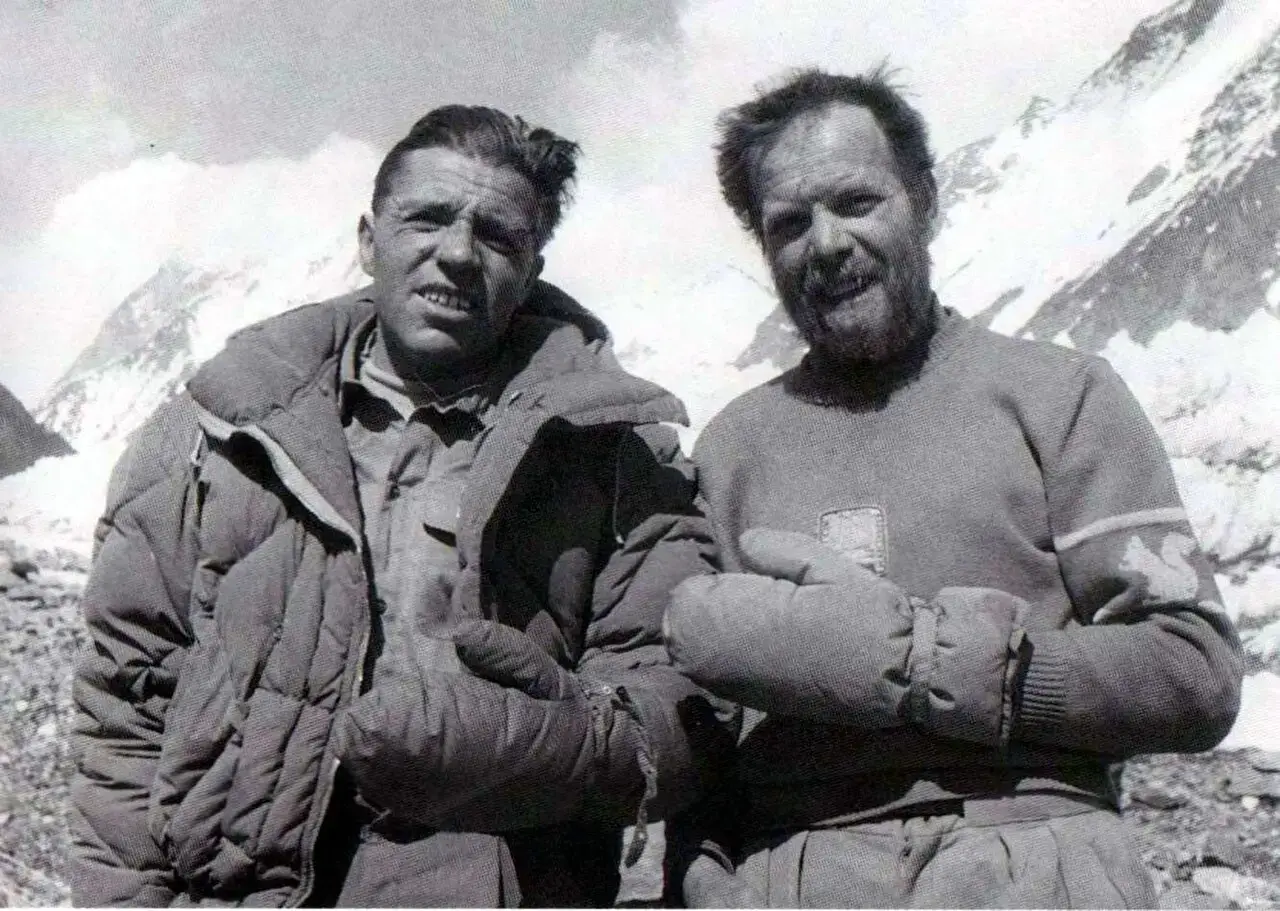 Historic black-and-white photo of Italian climbers Achille Compagnoni (left) and Lino Lacedelli (right) during the 1954 first ascent of K2. They stand arm-in-arm on a rocky, snowy slope with mountain peaks in the background; Compagnoni wears a quilted down jacket with a serious expression, while the bearded Lacedelli smiles slightly in a wool sweater with a squirrel patch and thick mittens.