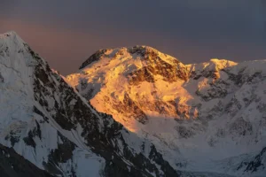 A dramatic mountain peak glows with intense golden-orange alpenglow during sunset or sunrise. The central summit, heavily snow-covered, catches warm sunlight on its rugged face and ridges, while surrounding peaks and deep valleys remain in cool blue shadows. The sky above transitions from deep twilight to soft orange near the horizon, highlighting the massive scale and icy texture of the high-altitude landscape.