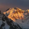 A dramatic mountain peak glows with intense golden-orange alpenglow during sunset or sunrise. The central summit, heavily snow-covered, catches warm sunlight on its rugged face and ridges, while surrounding peaks and deep valleys remain in cool blue shadows. The sky above transitions from deep twilight to soft orange near the horizon, highlighting the massive scale and icy texture of the high-altitude landscape.