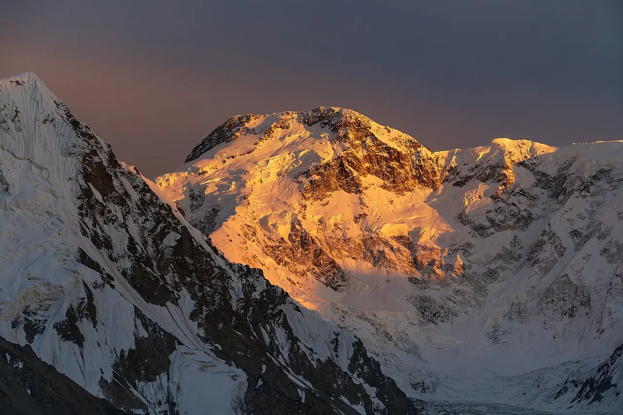 A dramatic mountain peak glows with intense golden-orange alpenglow during sunset or sunrise. The central summit, heavily snow-covered, catches warm sunlight on its rugged face and ridges, while surrounding peaks and deep valleys remain in cool blue shadows. The sky above transitions from deep twilight to soft orange near the horizon, highlighting the massive scale and icy texture of the high-altitude landscape.