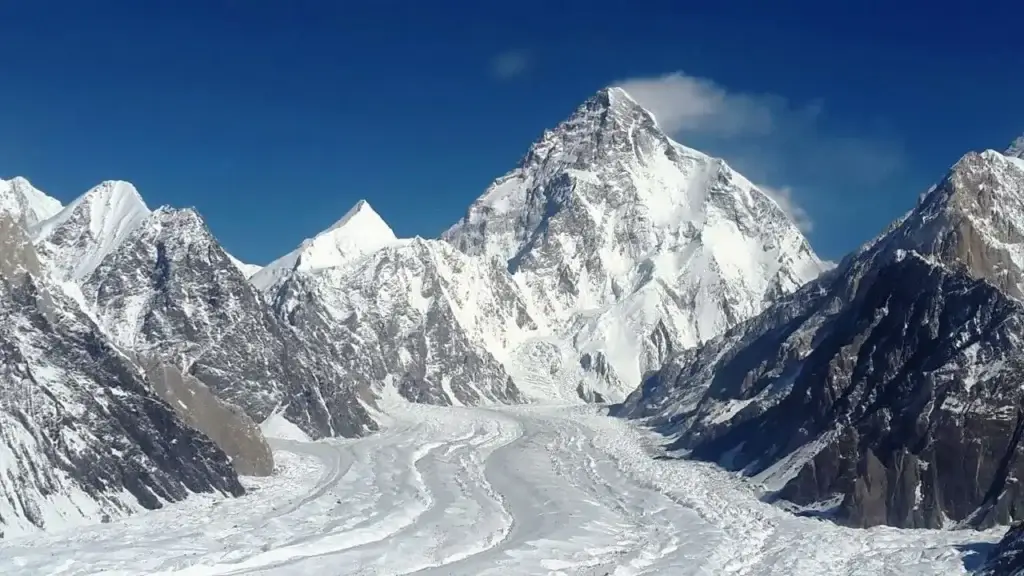 K2 (also known as Mount Godwin-Austen or Chhogori), the second-highest mountain in the world, rising sharply to its pyramidal summit under a clear blue sky. The heavily crevassed Godwin-Austen Glacier flows through the foreground, flanked by rugged, snow-covered peaks of the Karakoram Range.