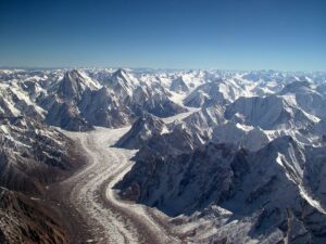 aAerial view of a massive glacier winding through the rugged Karakoram mountain range. A long, sinuous river of snow and ice flows down the center of the frame, flanked by steep, jagged peaks and ridges blanketed in snow. The glacier curves gently, surrounded by countless sharp, snow-covered summits stretching to the horizon under a clear deep-blue sky.