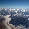 aAerial view of a massive glacier winding through the rugged Karakoram mountain range. A long, sinuous river of snow and ice flows down the center of the frame, flanked by steep, jagged peaks and ridges blanketed in snow. The glacier curves gently, surrounded by countless sharp, snow-covered summits stretching to the horizon under a clear deep-blue sky.