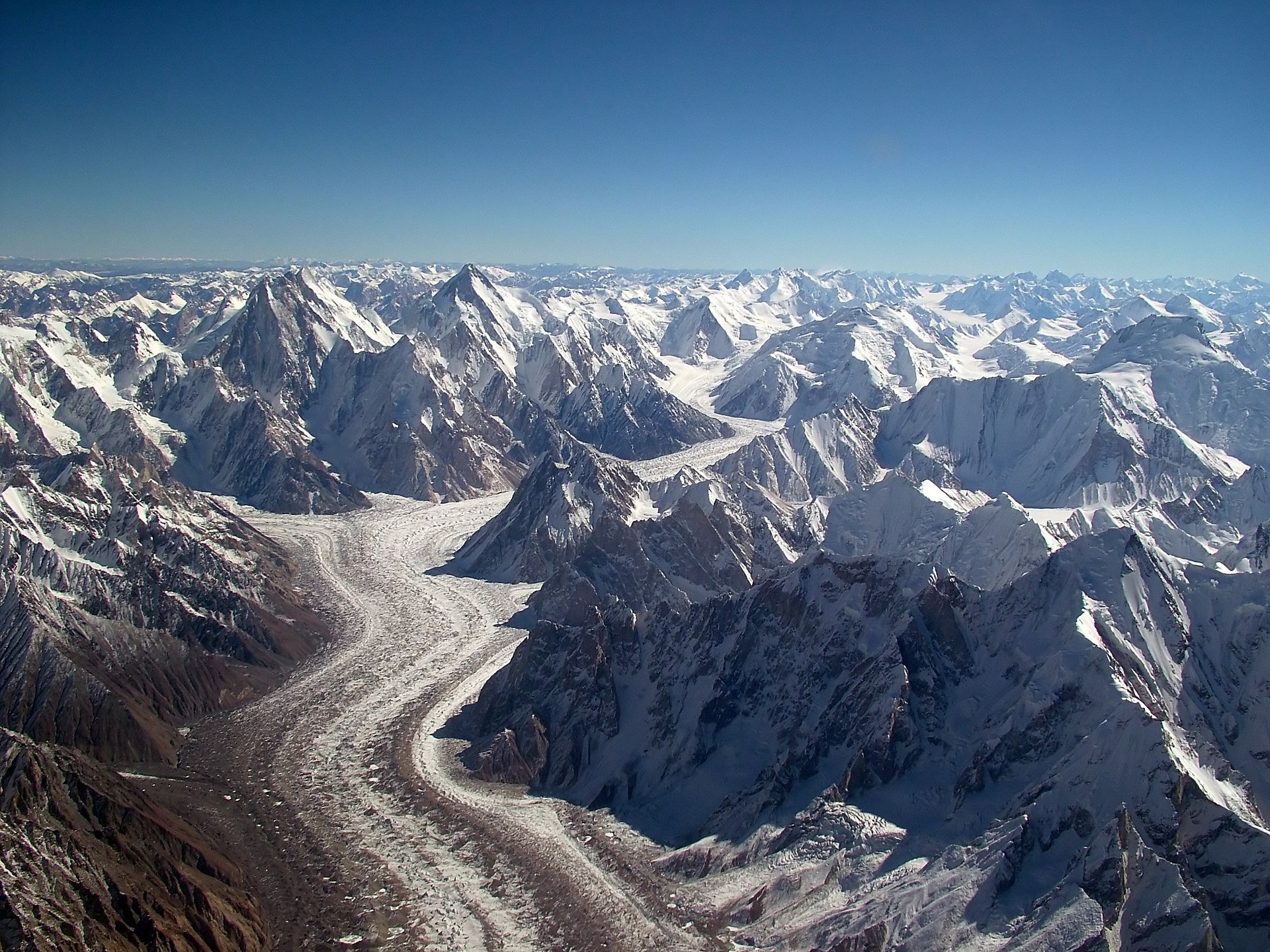 aAerial view of a massive glacier winding through the rugged Karakoram mountain range. A long, sinuous river of snow and ice flows down the center of the frame, flanked by steep, jagged peaks and ridges blanketed in snow. The glacier curves gently, surrounded by countless sharp, snow-covered summits stretching to the horizon under a clear deep-blue sky.