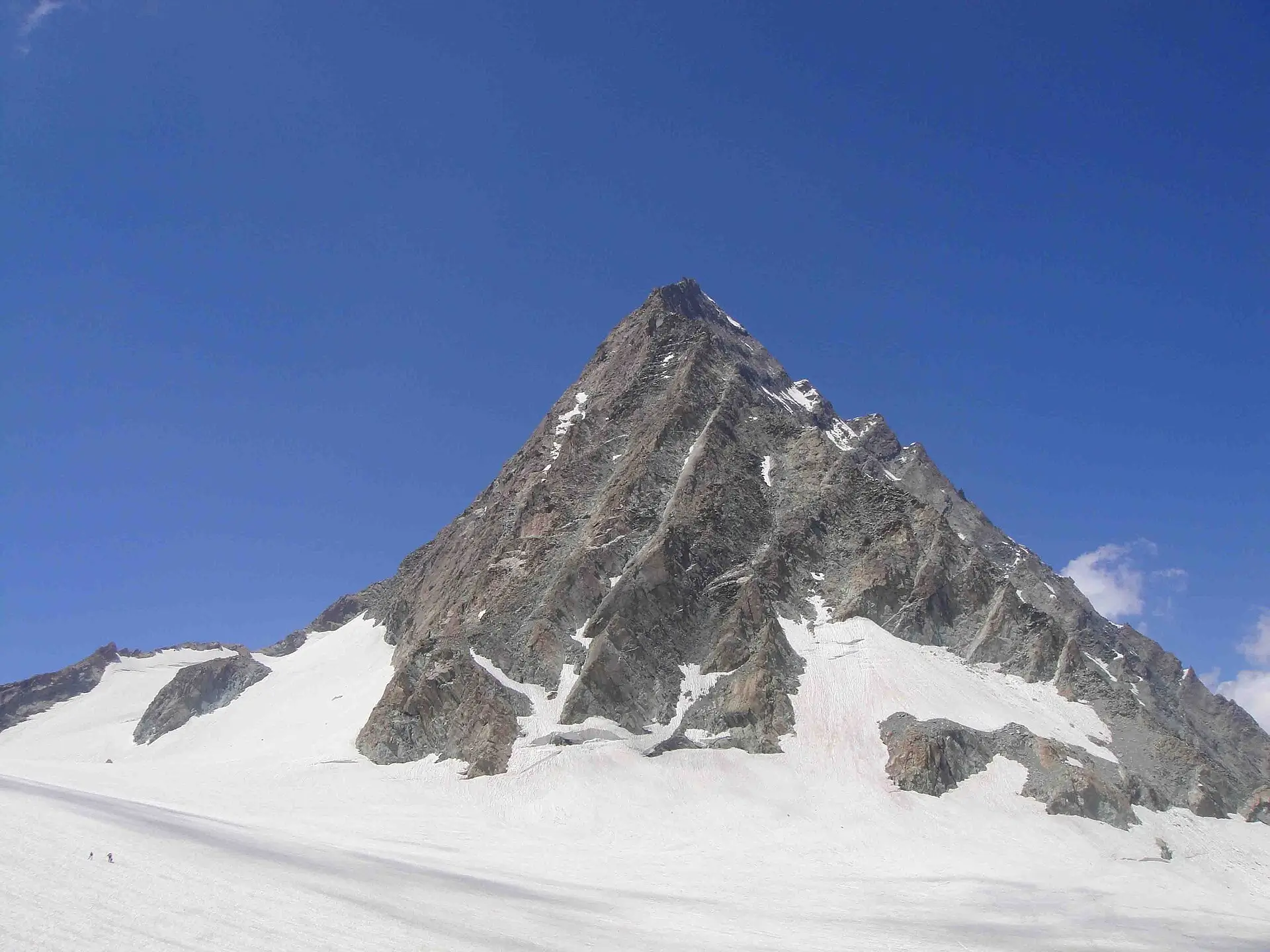 A dramatic, sharply pointed rocky mountain peak covered in patches of snow and ice, rising steeply from a wide glacier field under a clear deep blue sky. Tiny human figures (climbers) are visible on the snowy slope near the base, emphasizing the immense scale and high-altitude environment.