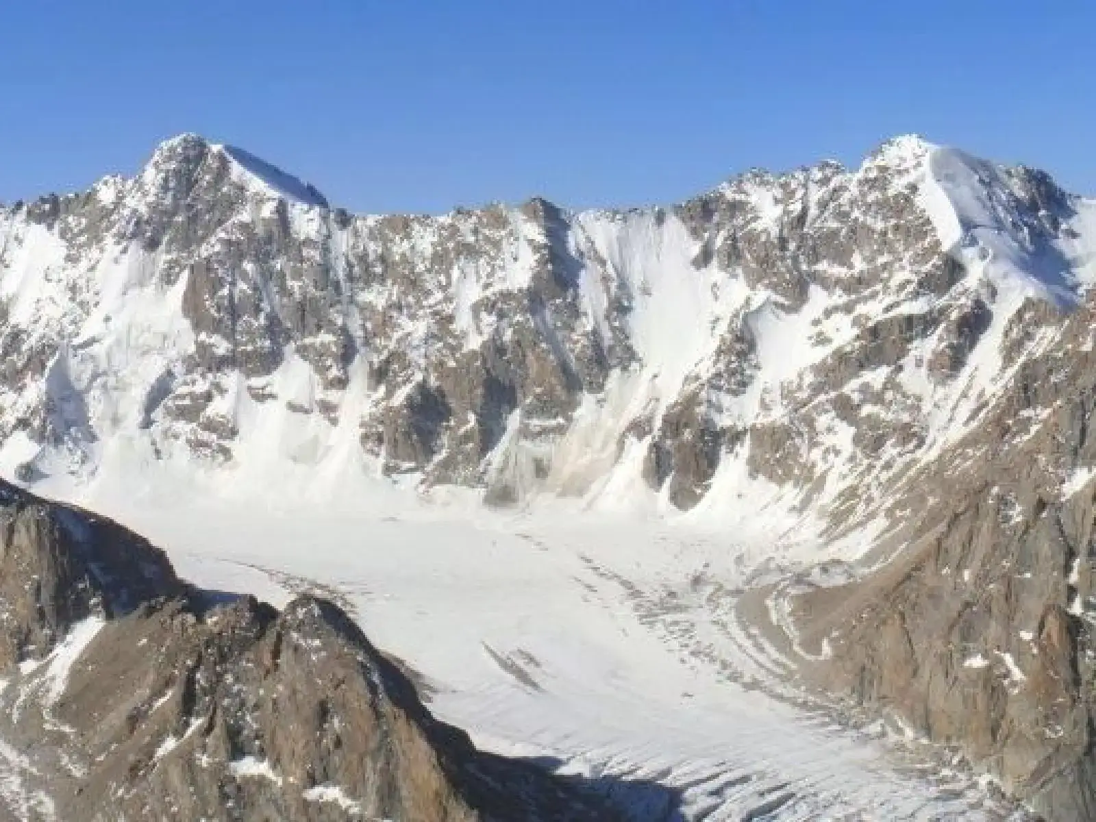 Expansive glacier flowing between rugged, snow-covered mountain peaks under a clear blue sky, with rocky ridges and ice fields visible in this high-altitude alpine landscape.