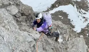A determined female mountaineer climbs a steep, rugged rock face in a high alpine environment. She wears a purple long-sleeve top, black pants, a black-and-white helmet, and climbing gear including a large white backpack with gear attached. She grips an ice axe in her right hand, which is planted into the rock, while her left hand holds the rock for balance. Her body is angled diagonally across the steep slab, with one leg extended and cramponed boot pressing against the granite. Snow patches cover parts of the surrounding rocks and the background shows a snowy mountain slope under bright daylight conditions.