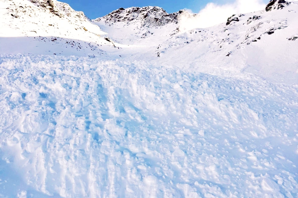 A wide view of a pristine alpine snowfield under a clear blue sky with some wispy clouds. Deep, wind-sculpted snow covers the ground, forming smooth, flowing drifts and wave-like patterns. In the background rise sharp, snow-covered mountain ridges and rocky peaks, with sunlight creating bright highlights and soft blue shadows in the snow.