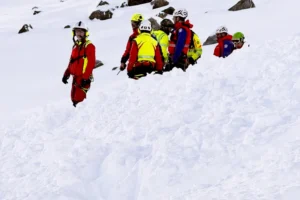 A mountain rescue team of eight people wearing brightly colored jackets (red, yellow, blue, green), helmets, and safety harnesses carefully navigates a steep, deep snow slope. They are moving in a line, some partially sunk into the snow up to their waists, with one rescuer in a red suit standing higher up looking toward the camera. Snowy rocky peaks are visible in the background.