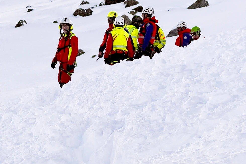 A mountain rescue team of eight people wearing brightly colored jackets (red, yellow, blue, green), helmets, and safety harnesses carefully navigates a steep, deep snow slope. They are moving in a line, some partially sunk into the snow up to their waists, with one rescuer in a red suit standing higher up looking toward the camera. Snowy rocky peaks are visible in the background.